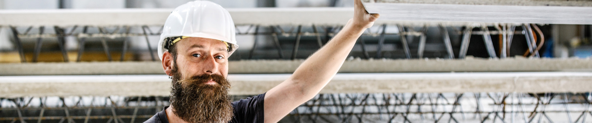 In einer Halle hält ein bärtiger Arbeiter mit weißem Helm und grauem T-Shirt eine lange Betonplatte am Kranhaken und stützt sie mit Werkzeug. Im Hintergrund stehen Maschinen, Regale und Materialien.