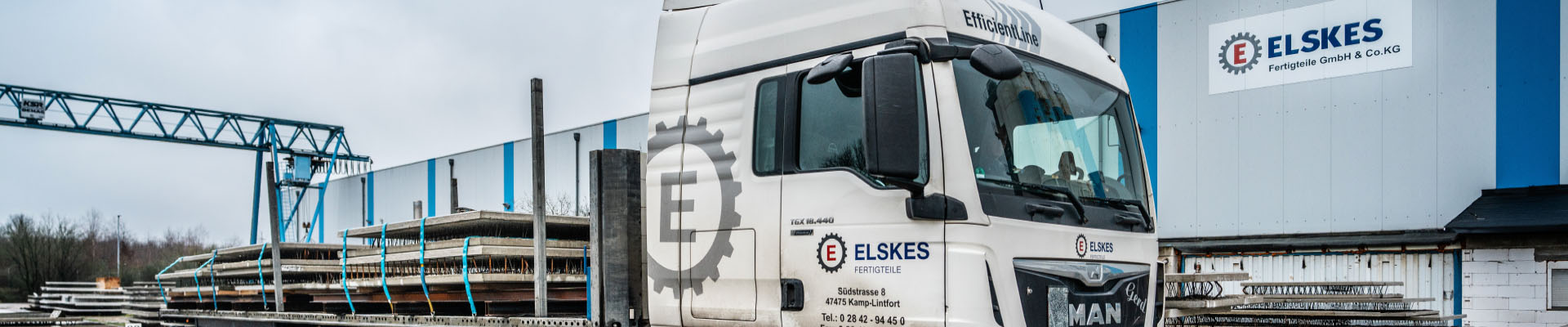 Ein weißer Lkw mit der Aufschrift „ELSKES“ steht vor einer blau weißen Industriehalle. Auf der Ladefläche liegen lange Metallplatten, gesichert mit türkisfarbenen Gurten. Im Hintergrund hebt ein Kran eine Platte.