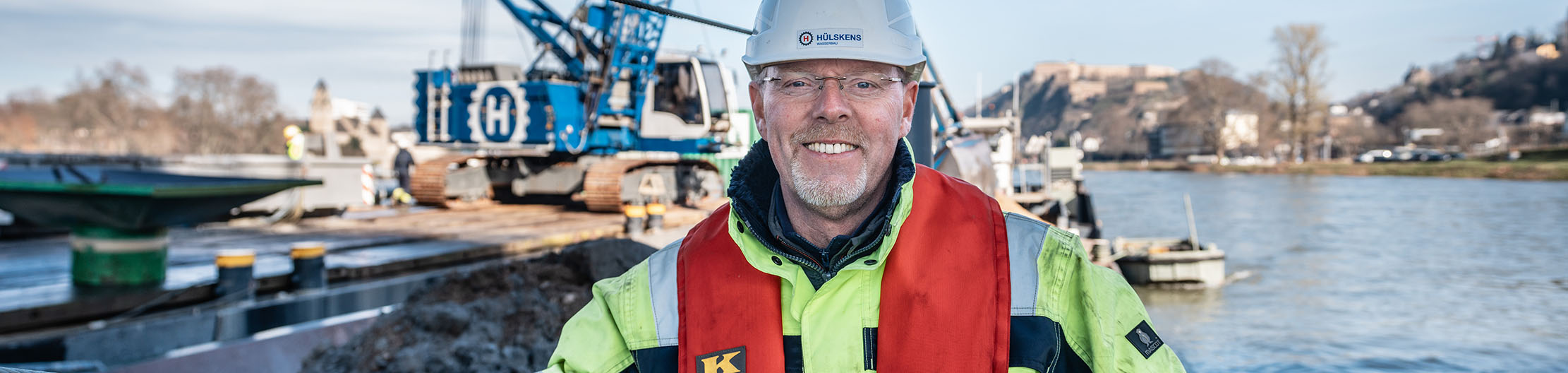 Person in Schutzkleidung mit Schutzhelm auf einer Baustelle am Wasser. Im Hintergrund sind eine Baumaschine auf einem Arbeitsponton, das Gewässer und das gegenüberliegende Ufer zu sehen.